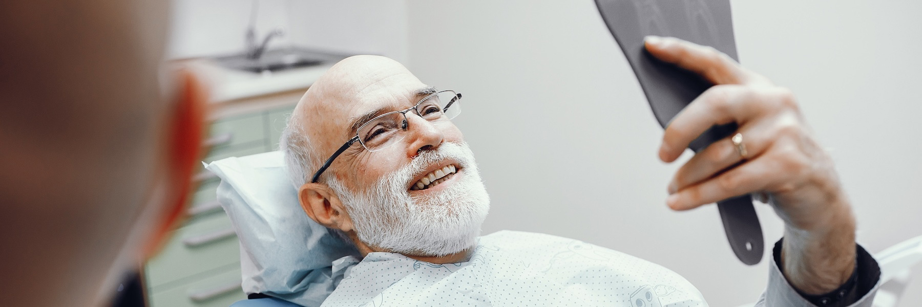 Smiling older man in dental chair viewing his teeth in a hand mirror held by dentist
